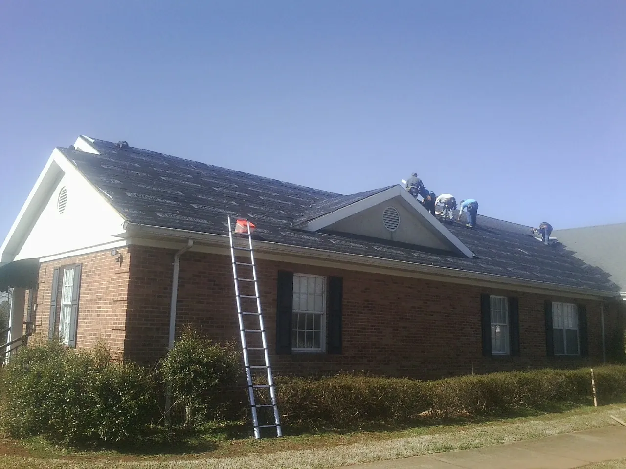Crew tearing shingles off a brick ranch roof with a ladder set — Emergency Roof Repair work by our crew in Flanagan