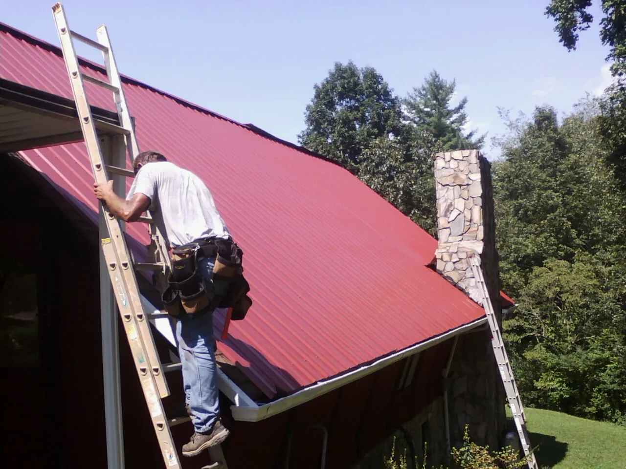 Roofer on a ladder beside a red metal roof and stone chimney — Soffit & Fascia Repair by Roofing Brothers in Flanagan